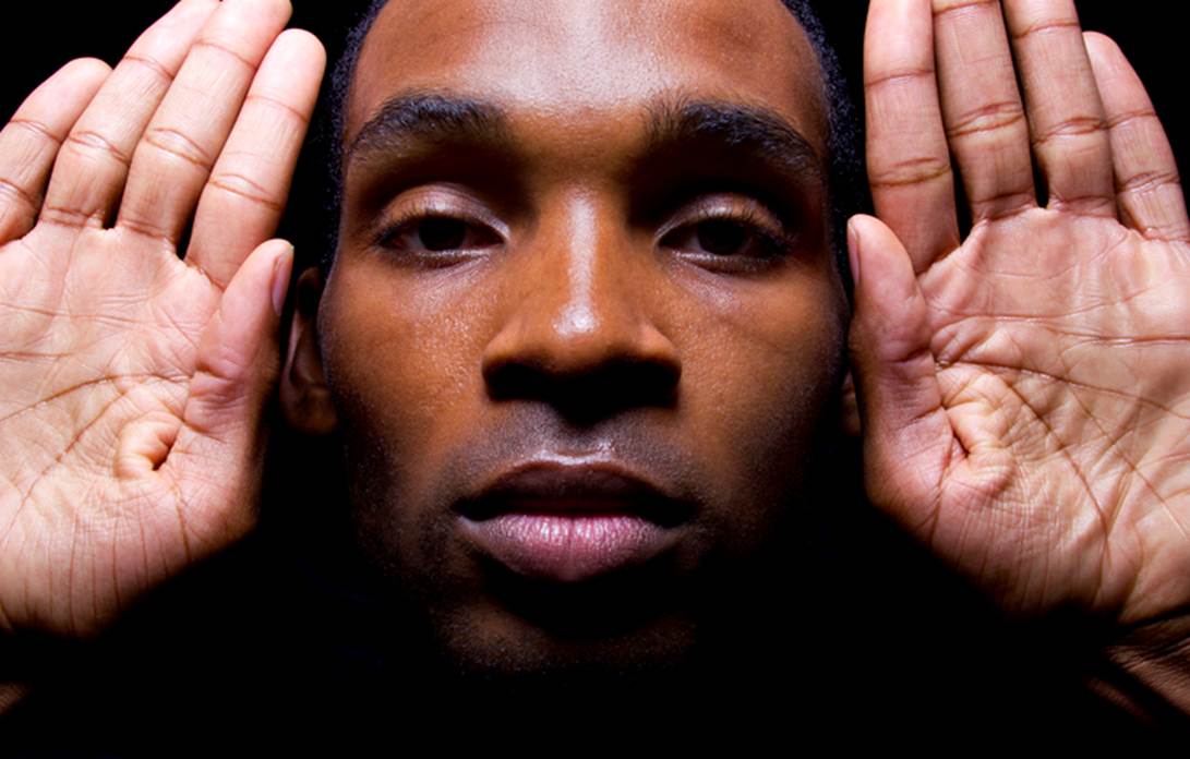 A close-up image of a man with his hand raised to the side of his face.